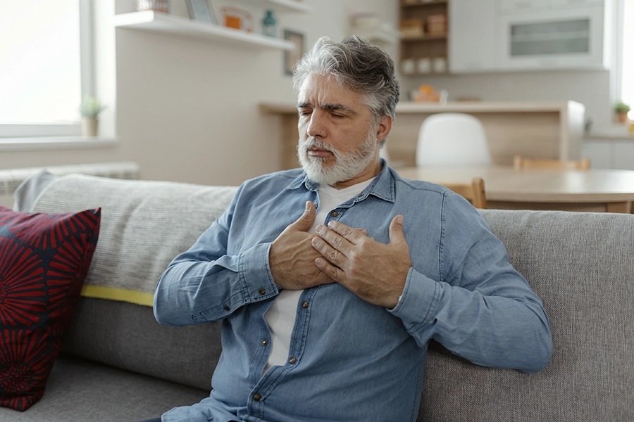Older man with a gray beard sits on a couch at home with both hands pressed to his chest, eyes closed in discomfort.