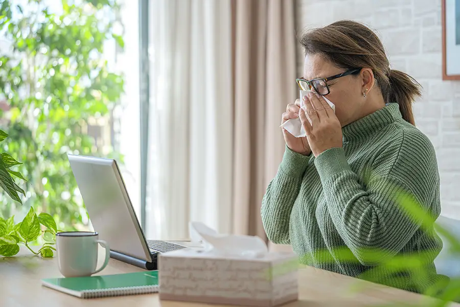 A woman sitting at a home desk blows her nose into a tissue while working on a laptop, with a tissue box nearby. She wears glasses and a green sweater, suggesting cold or allergy symptoms.